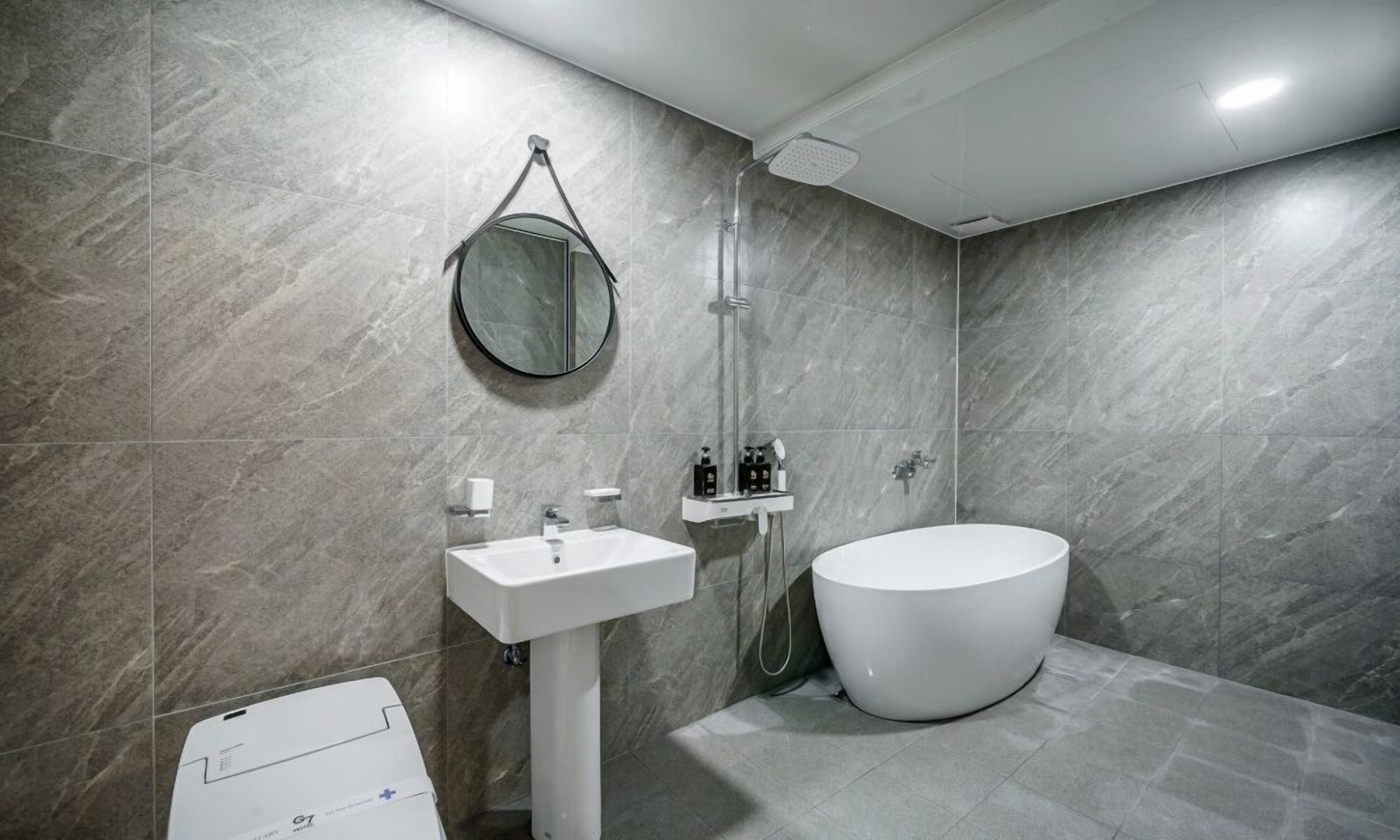 A modern bathroom with a freestanding bathtub, a pedestal sink, a handheld showerhead, and a round mirror.