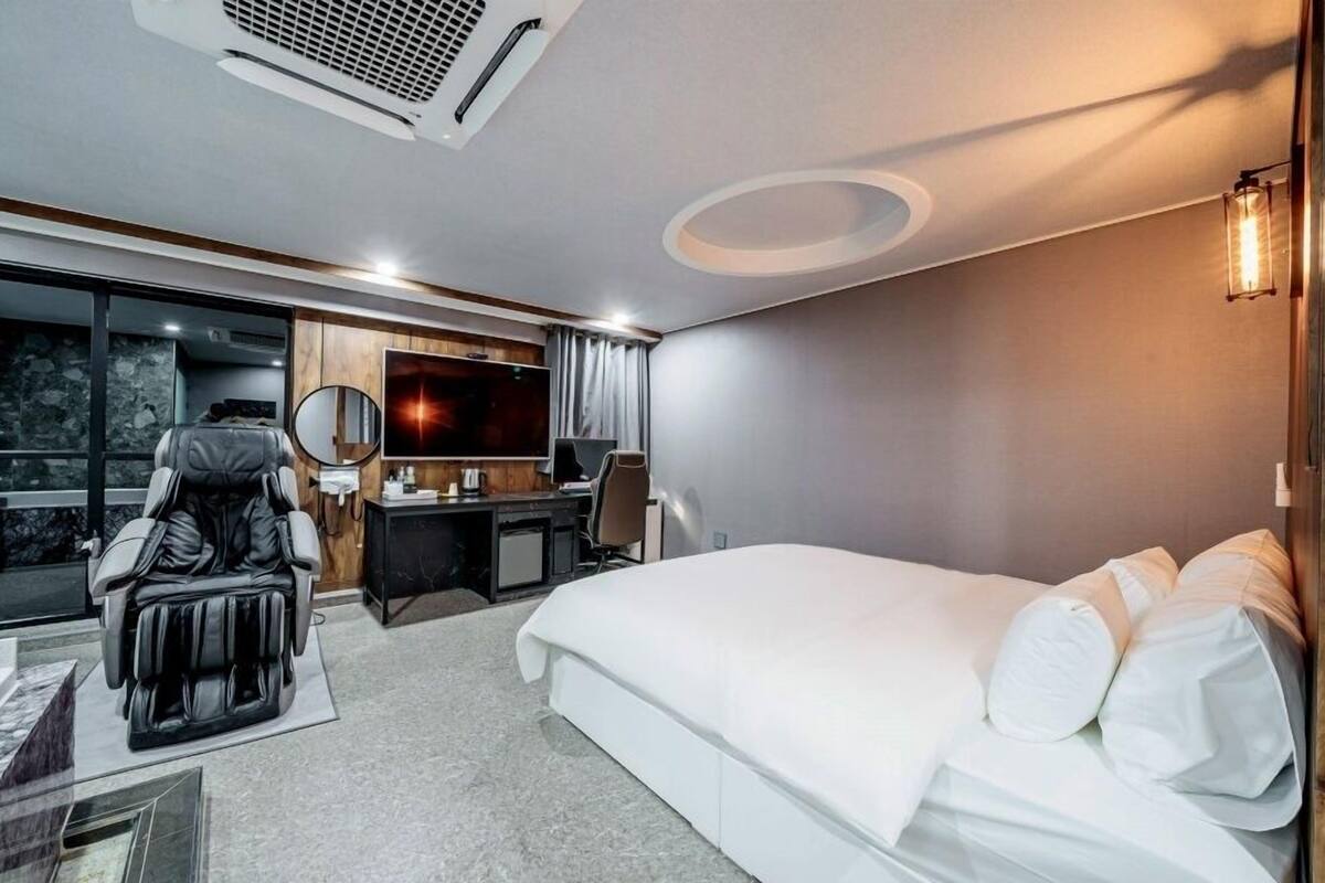 A modern hotel room with a large bed, a desk with a computer, a massage chair, and a large window.