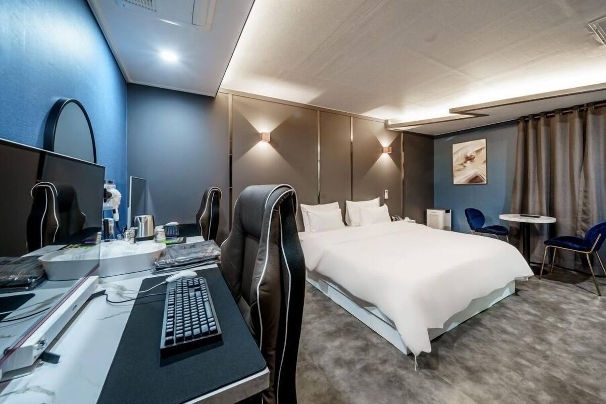 A modern hotel room with a large bed, a desk with a computer, a chair, and a small table.