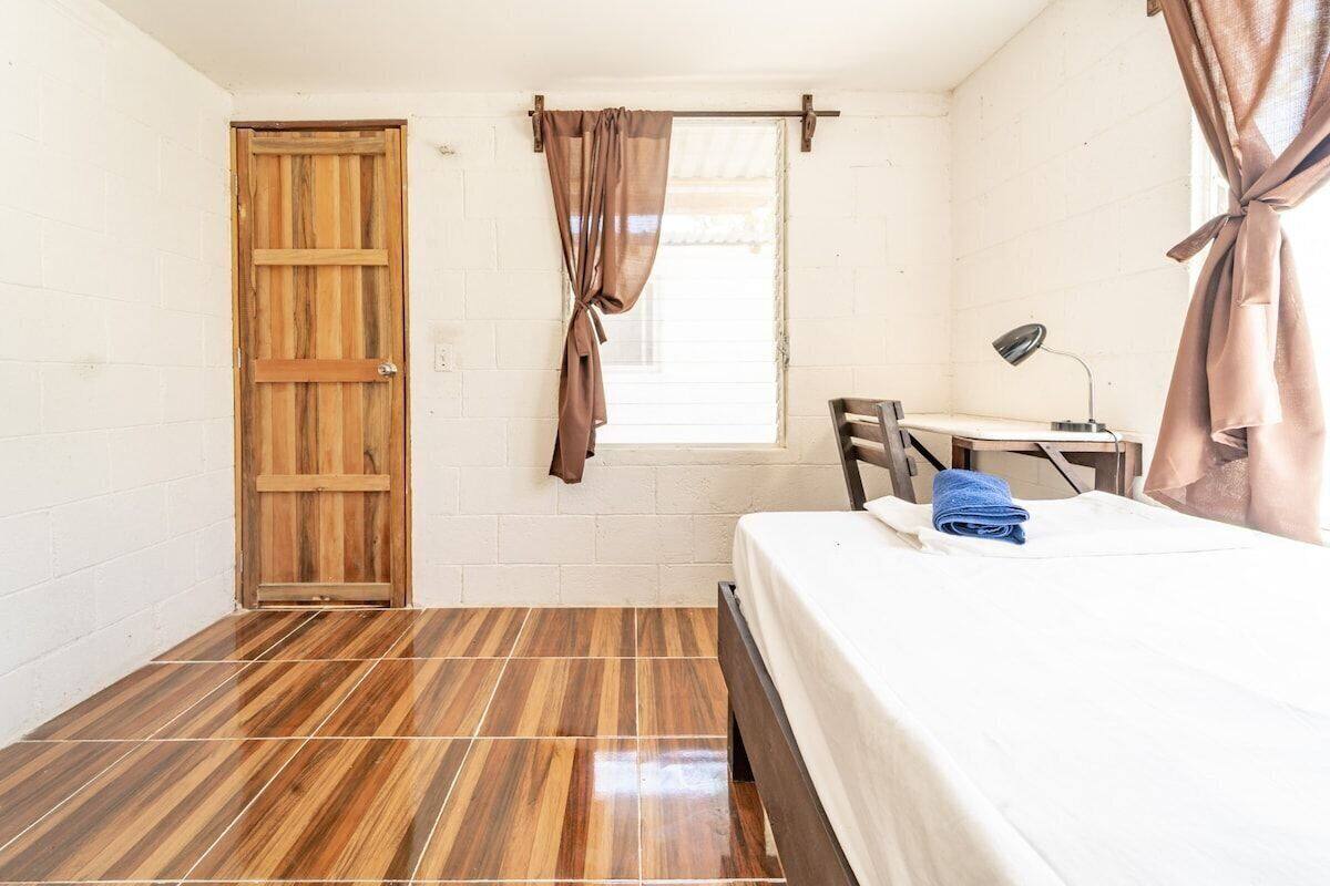 Una habitación pequeña y limpia con puerta de madera, cama con colchón blanco, escritorio con lámpara y ventana con cortinas.