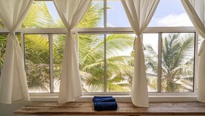 Quarto com uma grande janela, cortinas brancas, um banco de madeira e vista para palmeiras.