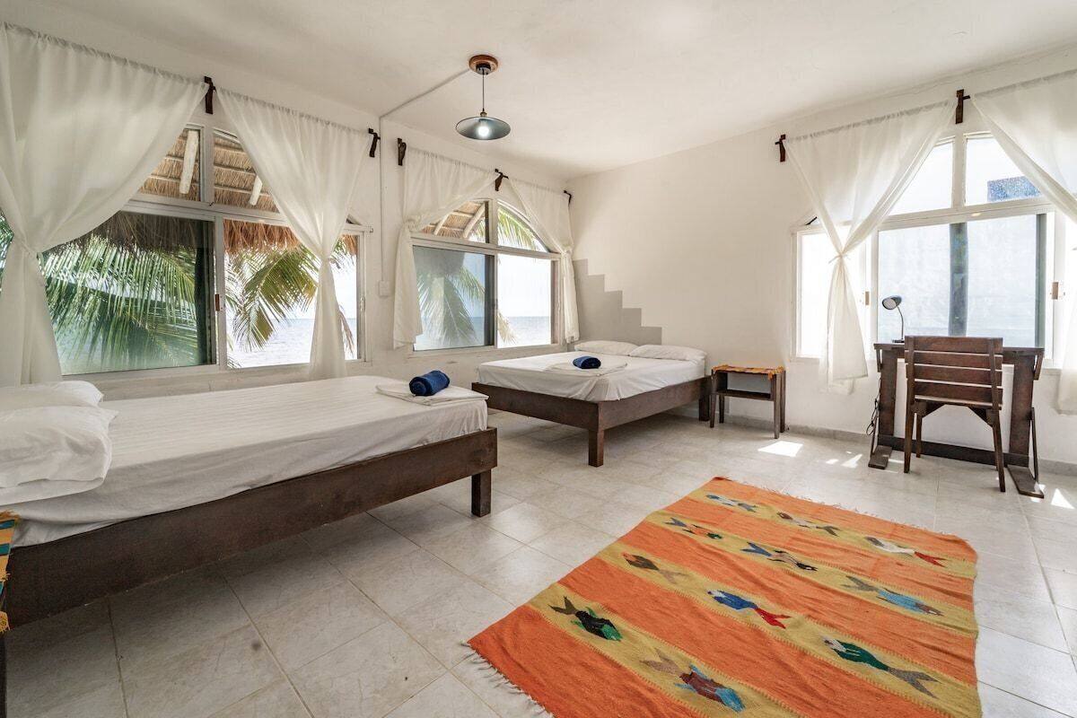 Una habitación con dos camas, una mesita, una silla y un ventanal con cortinas.