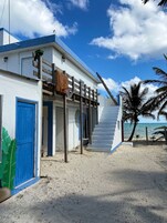 Una casa de playa con una puerta azul, una escalera que lleva a una terraza de madera y vistas al océano.