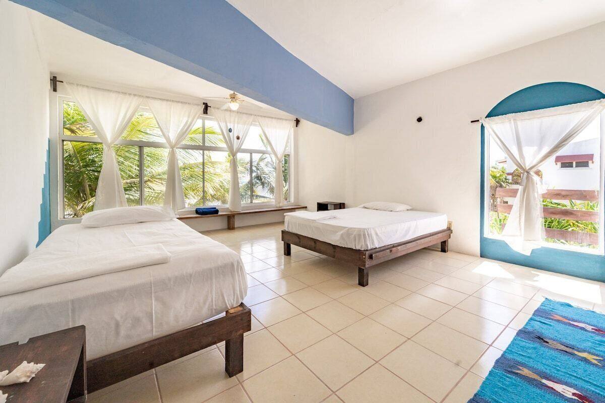 Una habitación con dos camas, ventanas amplias con cortinas, un ventilador de techo y vistas a la vegetación.