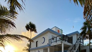 Um edifício à beira-mar com uma escadaria que leva a uma varanda, cercado por palmeiras e um mural na parede.