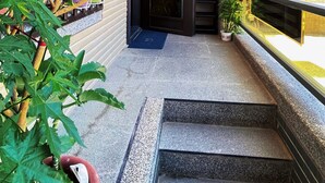 Una entrada de edificio con una puerta de madera, escalones que la conducen y plantas en macetas a la izquierda.