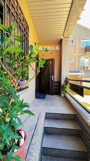 Stairs lead up to the entrance, flanked by potted plants and a balcony with a railing.