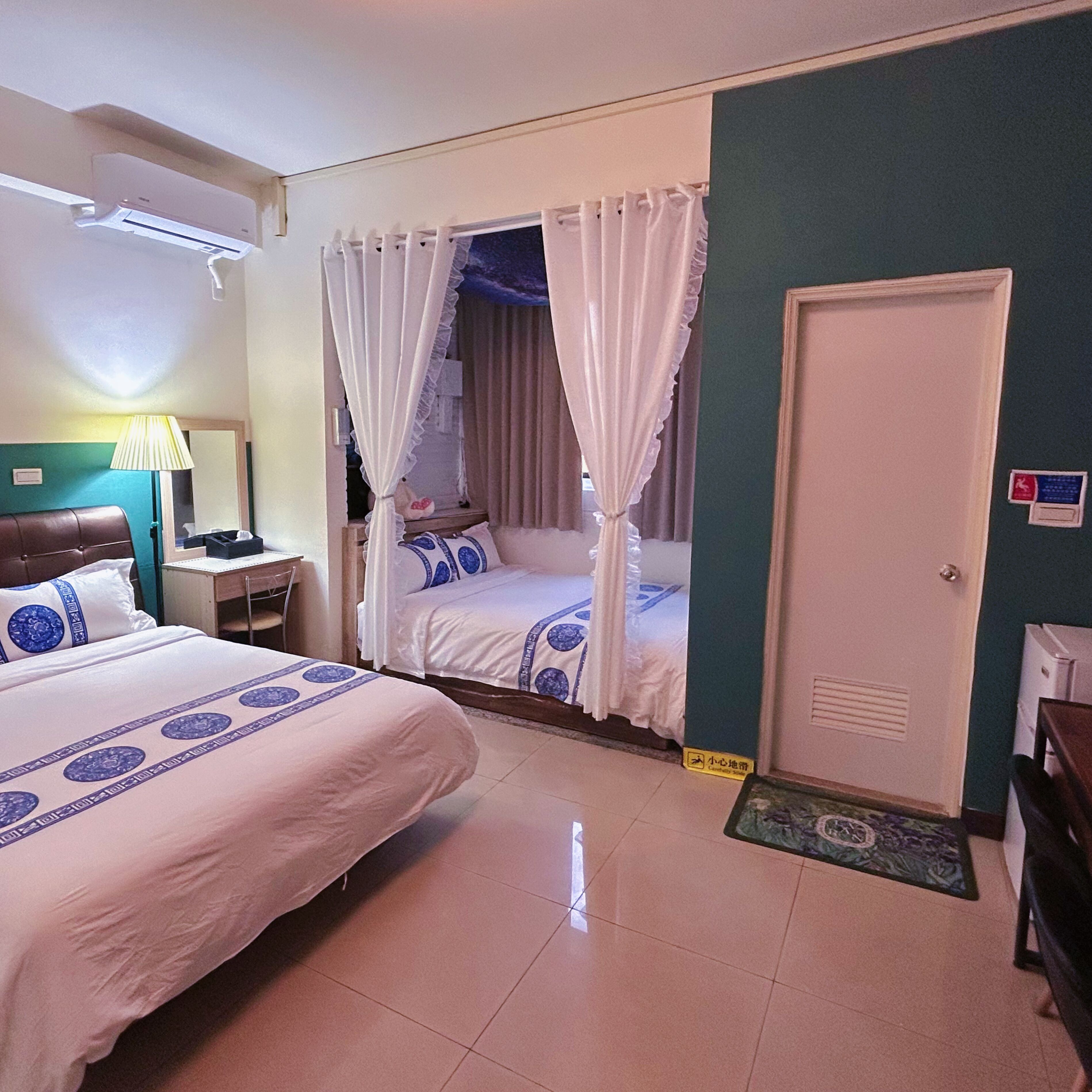A hotel room with two beds, a desk, a lamp, an air conditioning unit, and a door with a sign.