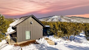 Una cabaña pequeña de un solo piso con techo inclinado, una puerta y una ventana. Está rodeada de nieve y árboles, con una montaña al fondo.
