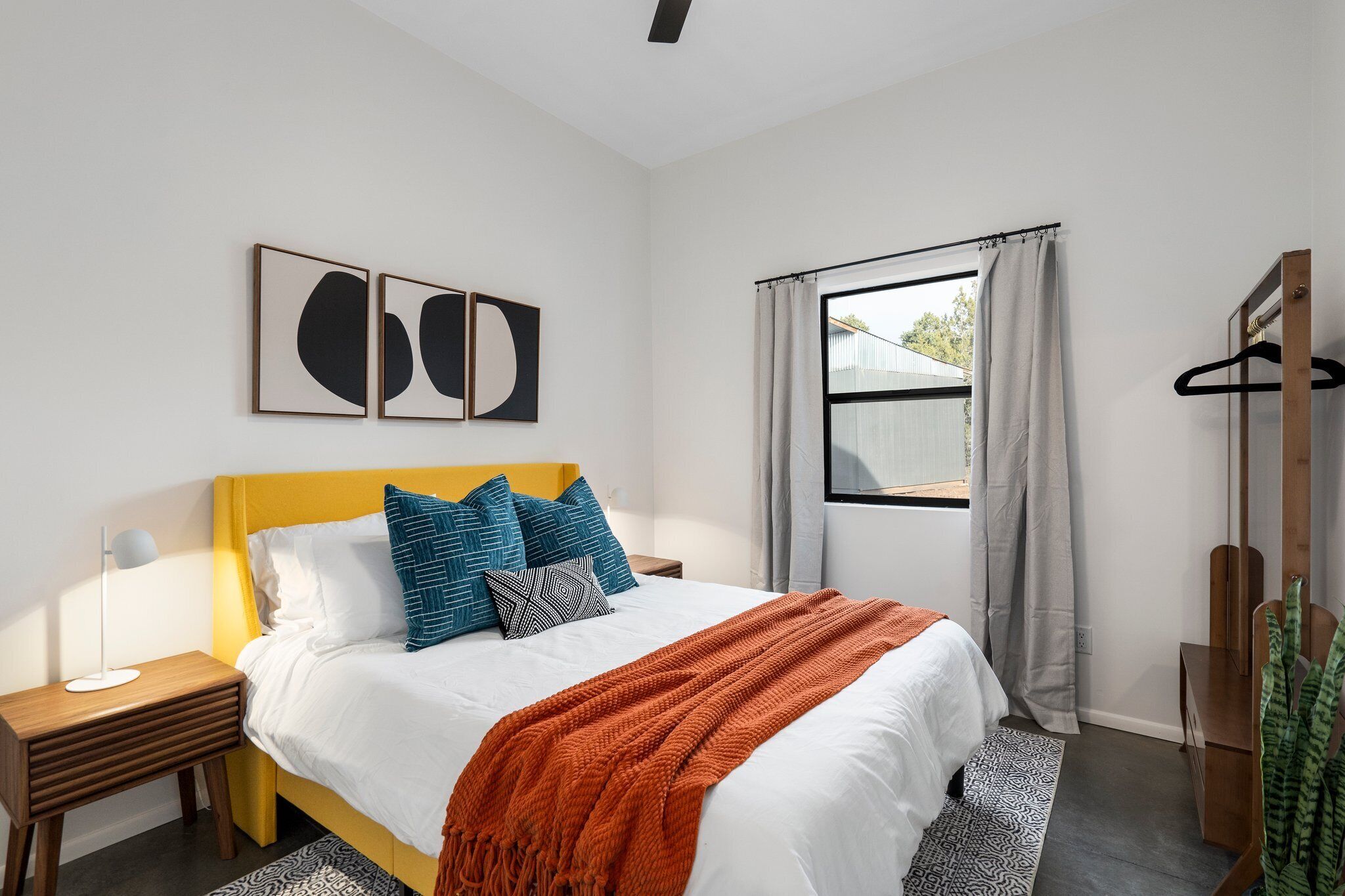 Un dormitorio moderno con cabecera amarilla, ropa de cama blanca y un ventanal con cortinas.