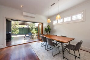 Uma ampla sala de jantar com uma mesa e cadeiras de madeira, uma janela grande com persianas e vista para o exterior.
