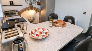 Et køkken med en stenovn, tostere, kaffemaskine og et spisebord dækket med brød, vin og en tallerken med blomster.