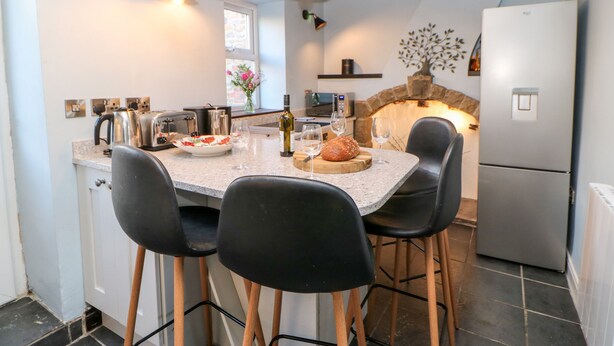 Et køkken med en central ø, sorte stole, et køleskab og en stensætning som pejs.