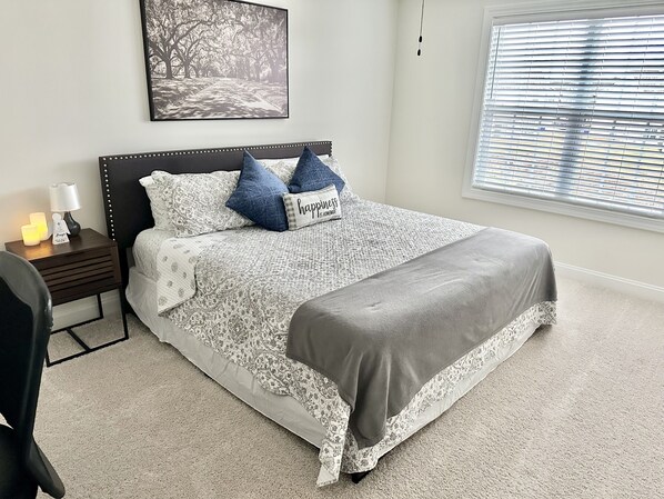 A bedroom with a bed, a nightstand, a lamp, a framed picture on the wall, and a window with blinds.