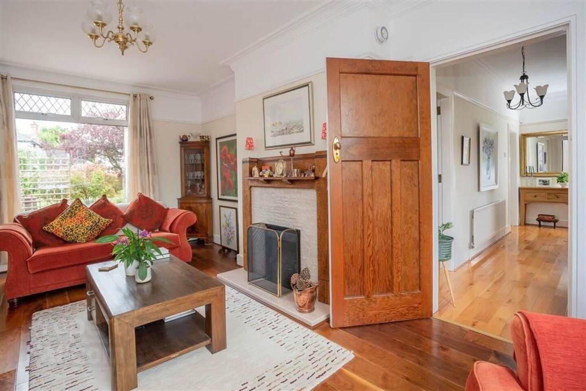 Un salon spacieux avec un canapé rouge, une table basse et une cheminée. Une porte en bois donne accès à une autre pièce, et un couloir mène à un autre espace, agrémenté d’un miroir et d’une plante.