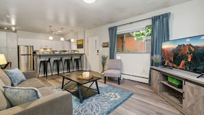 Un salon moderne avec un canapé, une table basse et un téléviseur. On y trouve également un coin cuisine avec des tabourets de bar et un réfrigérateur. Une fenêtre avec des rideaux est visible, et une plante est placée près de la fenêtre.
