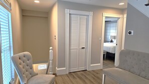 A hotel room with a large bed, a seating area with two chairs, a white wardrobe, and a view of the adjacent room.