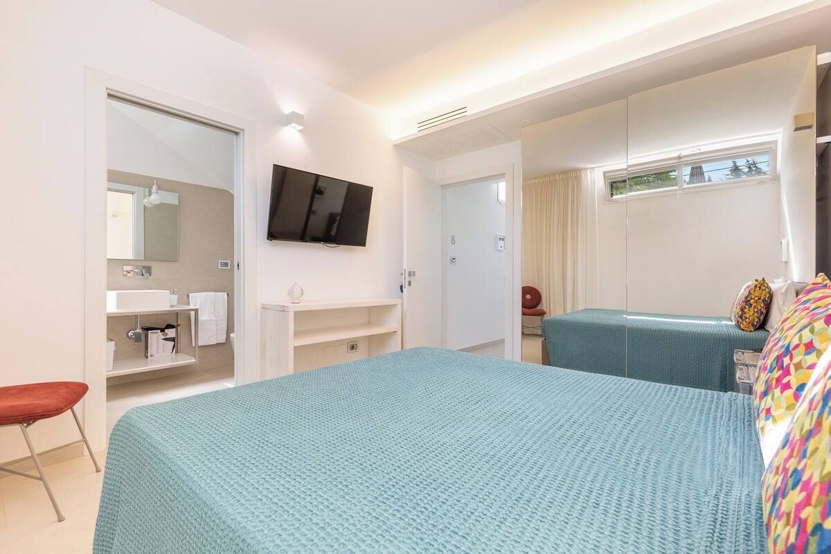 A hotel room with two beds, a TV, a bathroom with a mirror, and a window.