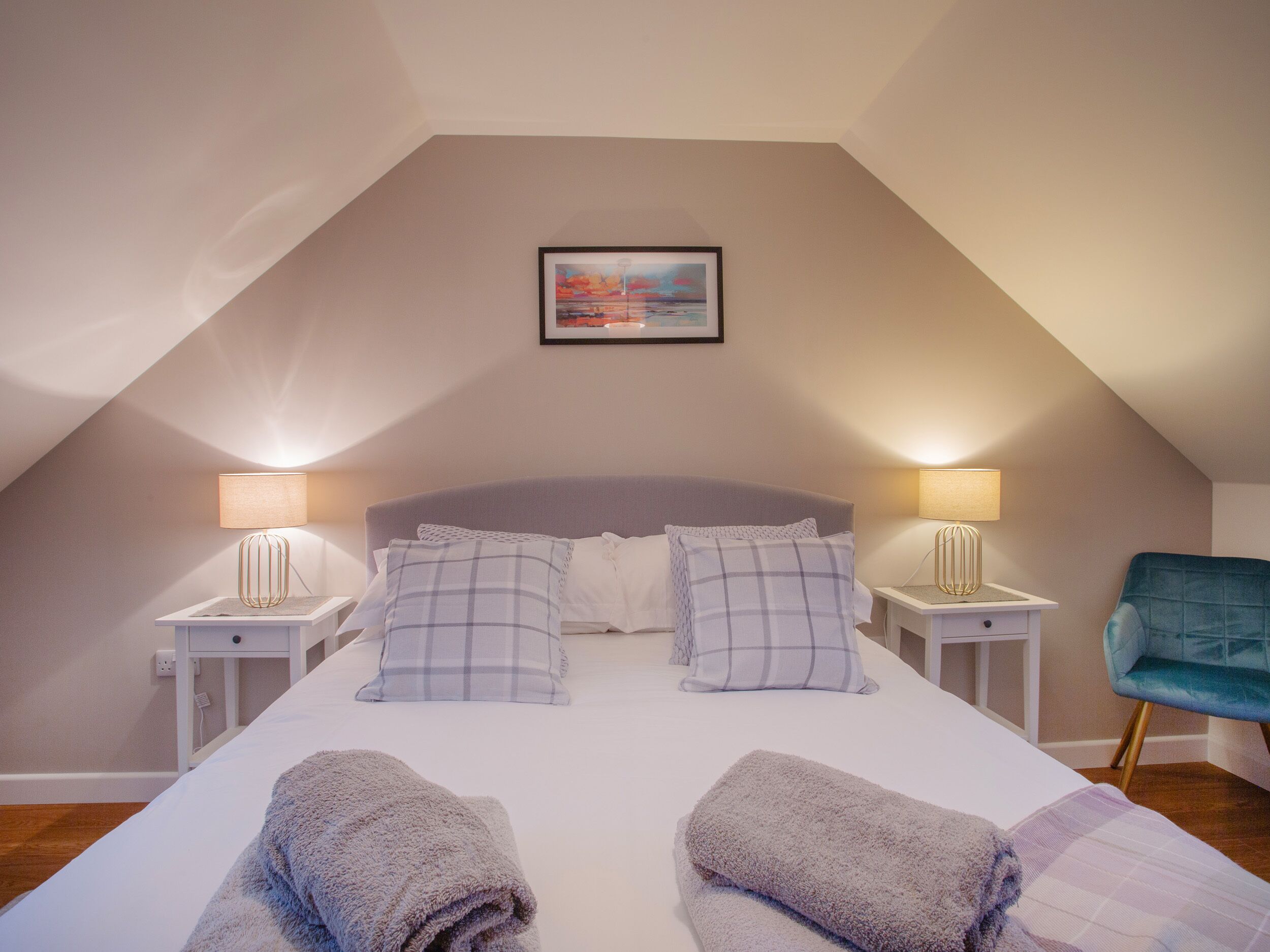 Een slaapkamer met een schuine kap, een bed met twee kussens, twee bedlampen, een stoel en een lijst met een foto aan de muur.