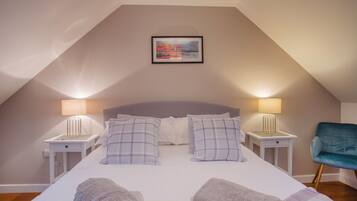 Een slaapkamer met een schuine kap, een bed met twee kussens, twee bedlampen, een stoel en een lijst met een foto aan de muur.