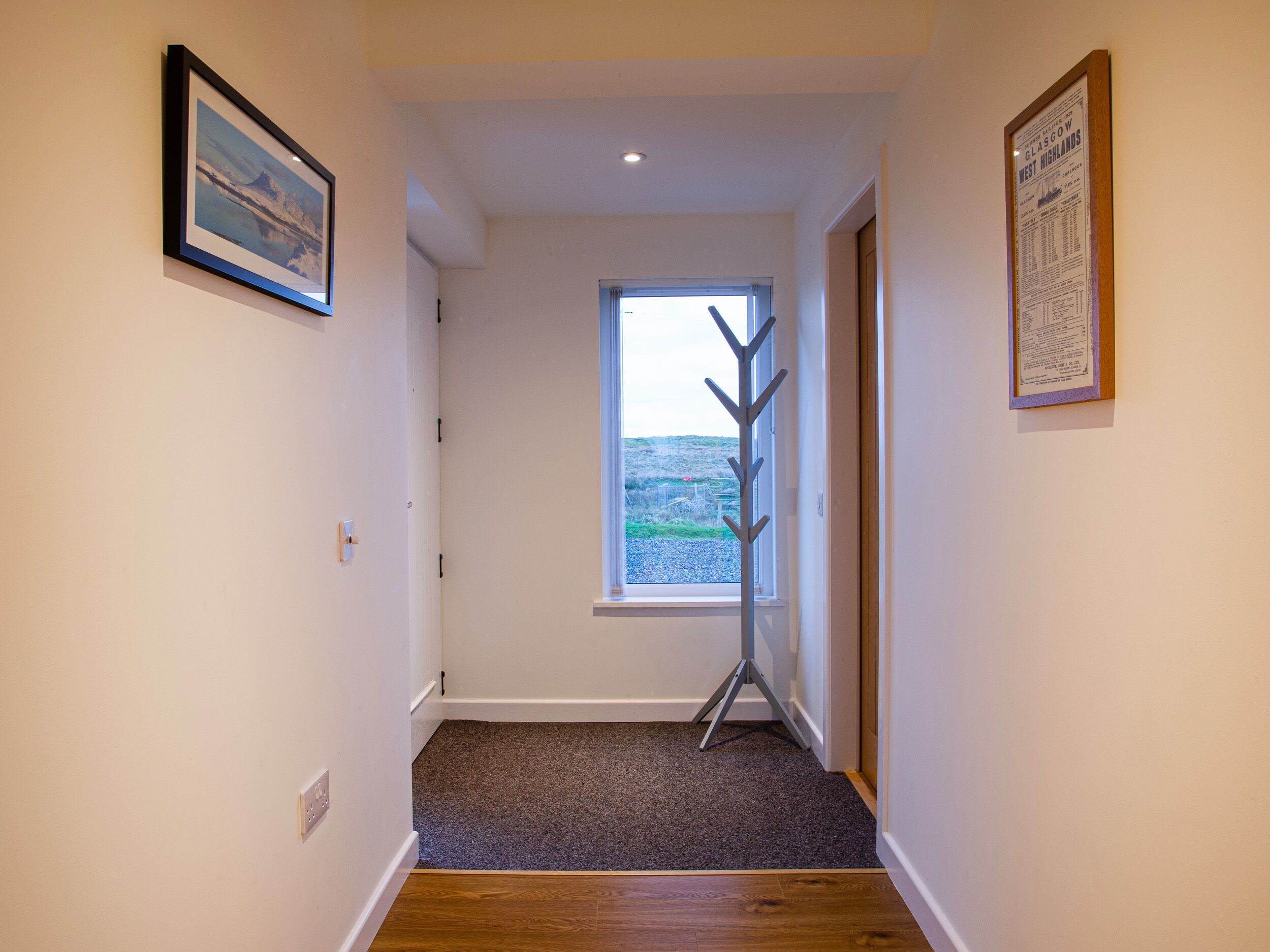 A hallway with a wooden floor, a window with a view of the countryside, and two framed pictures on the walls.