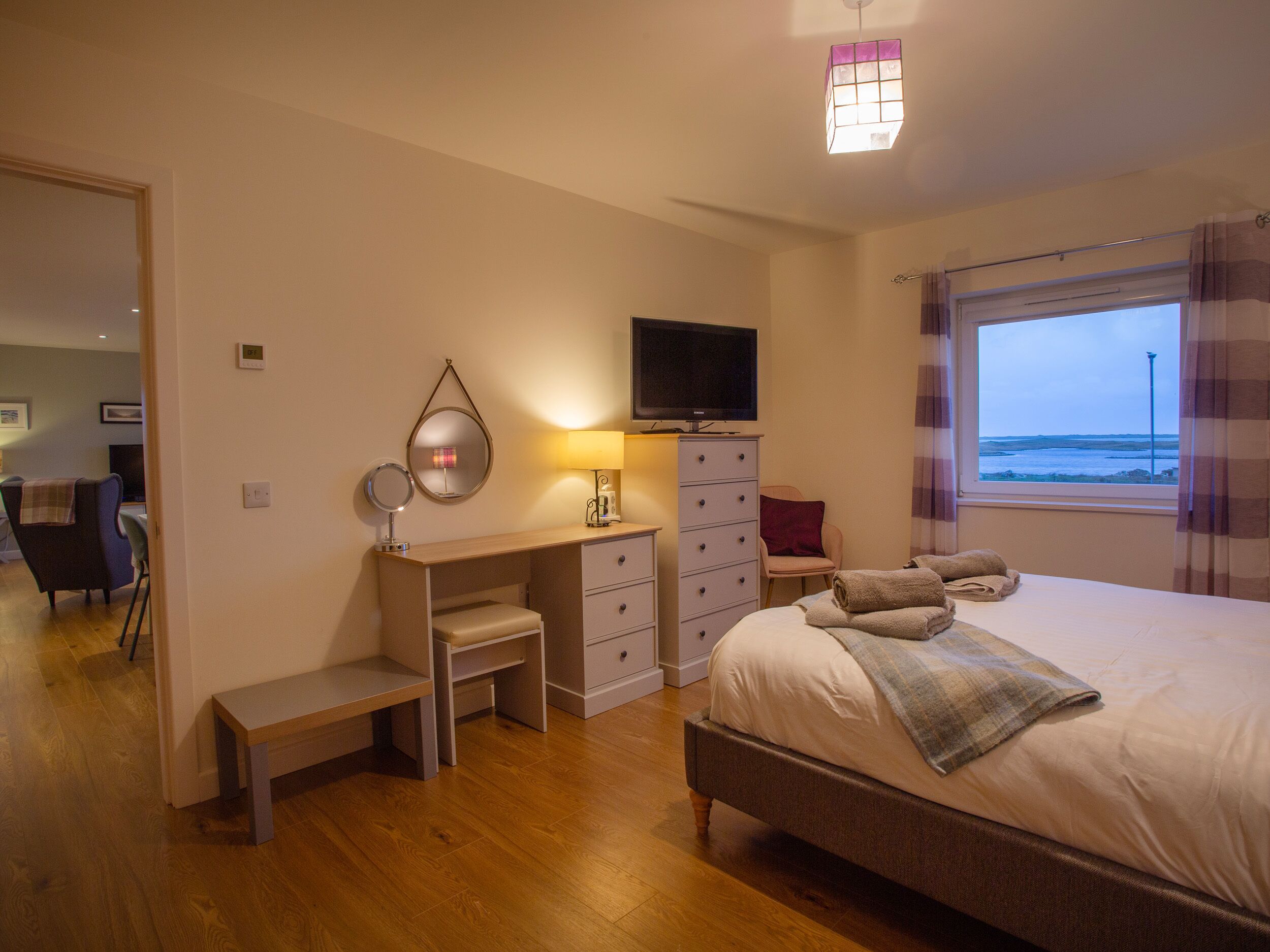 A bedroom with a bed, a desk, a chair, a television, and a window with a view of the sea.