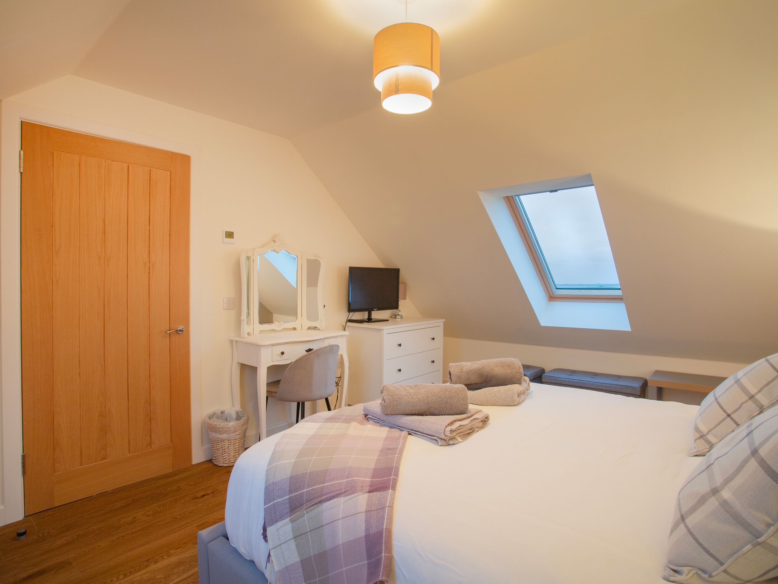 A bedroom with a sloped ceiling, a wooden door, a vanity with a mirror, a television, a bed with a plaid blanket, and a skylight.