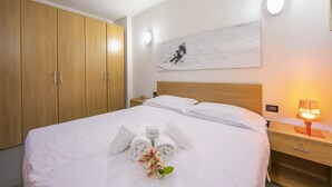 A neatly made bed with towels and a flower, a wooden wardrobe, a painting on the wall, and a bedside table with a lamp. - Teleo 104 Ski In - Ski Out 50m, Sestriere, Italy (Sestriere)