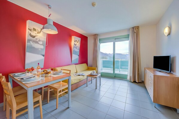 A modern living room with a red accent wall, a sofa, a dining table with chairs, a TV, and a large window with a view of mountains. - Teleo 105 Ski In - Ski Out 50m, Sestriere, Italy (Sestriere)