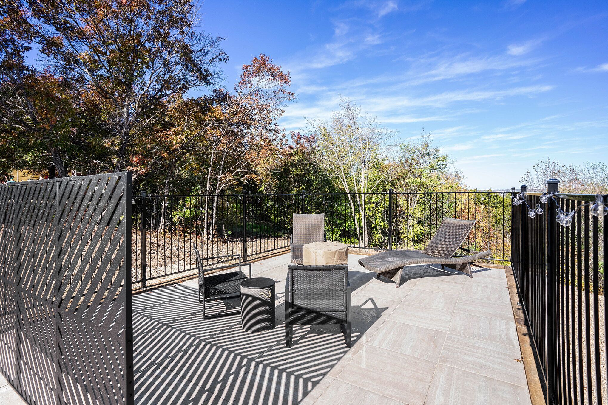 An outdoor patio with a black metal fence, two wicker chairs, a small round table, and a lounge chair.