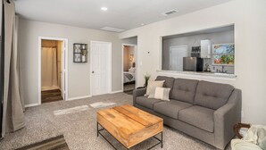 A living room with a grey sofa, a wooden coffee table, and a kitchen visible through an open doorway. - The Southern Sapphire: A Cozy Lake View Getaway (Granbury)