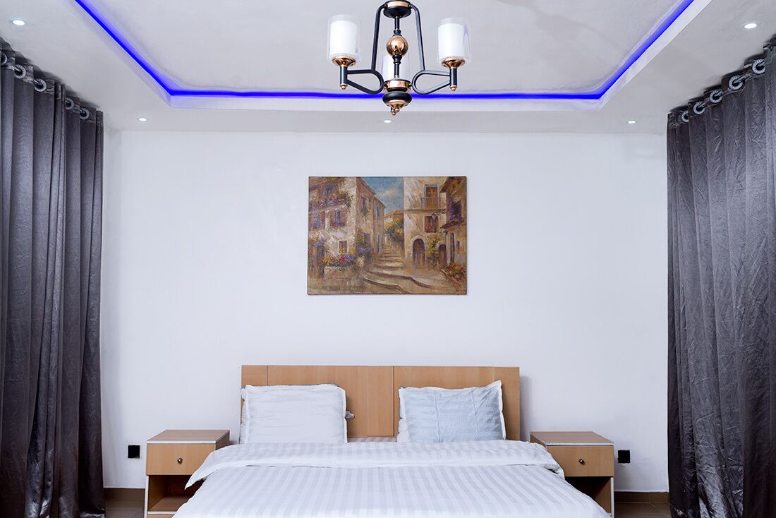 A modern bedroom with a large bed, two bedside tables, a painting on the wall, and a ceiling light fixture.