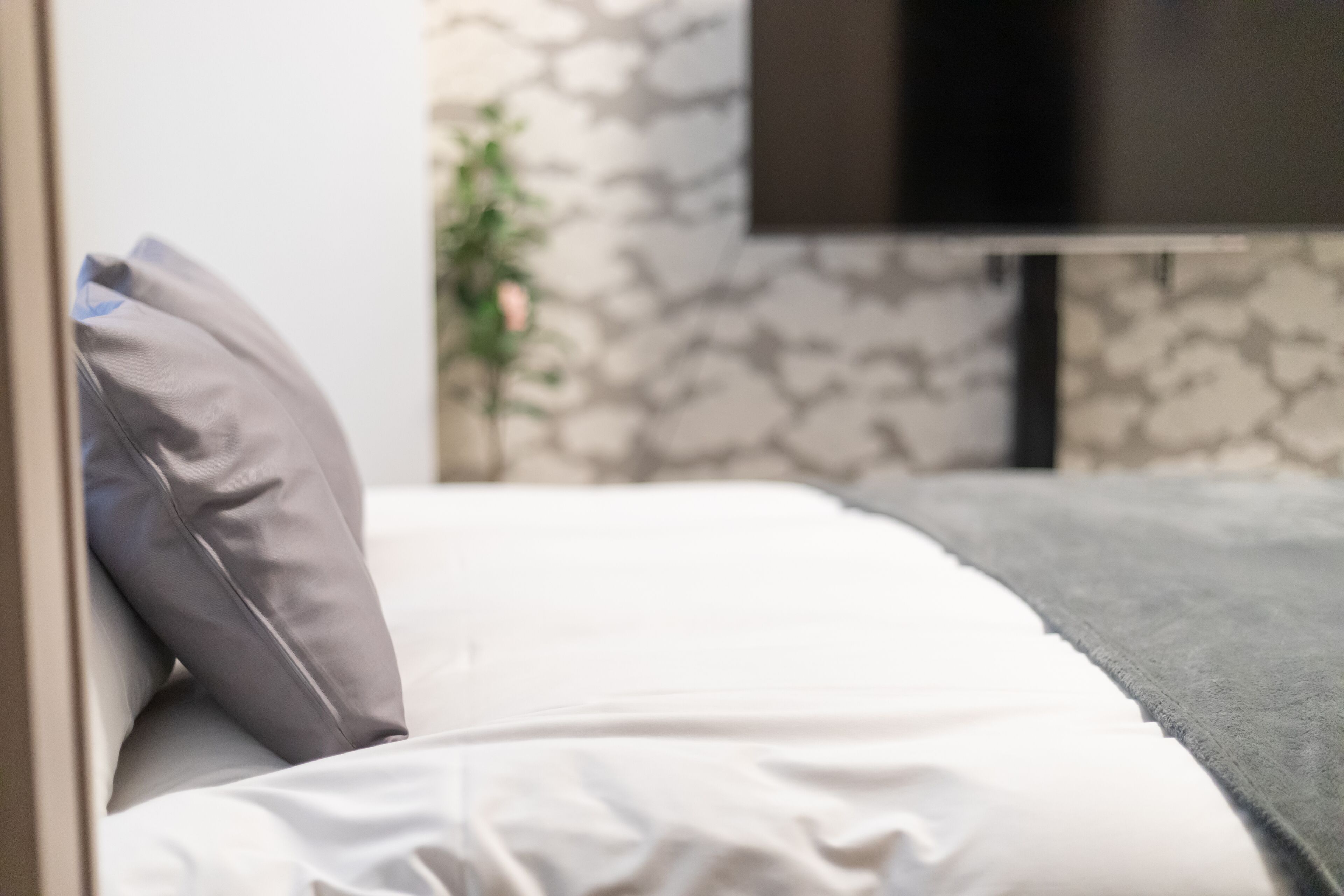 A bed with white bedding and a grey pillow, a grey blanket, and a television mounted on the wall.