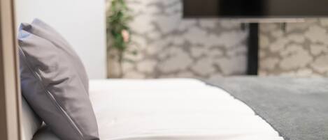 A bed with white bedding and a grey pillow, a grey blanket, and a television mounted on the wall.