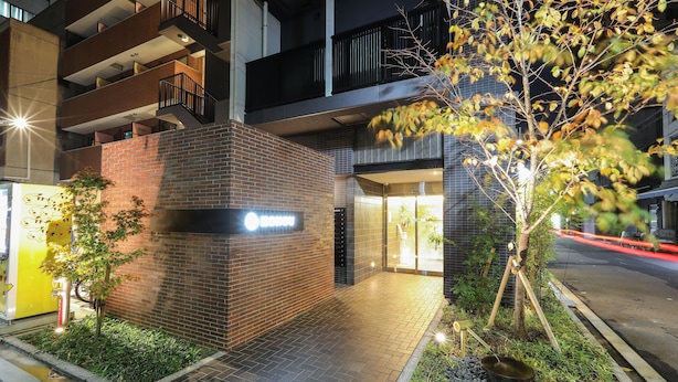 Un edificio moderno con facciata in mattoni, una insegna che indica un'attività commerciale o un servizio, e un piccolo giardino con un albero e una ciotola in metallo.