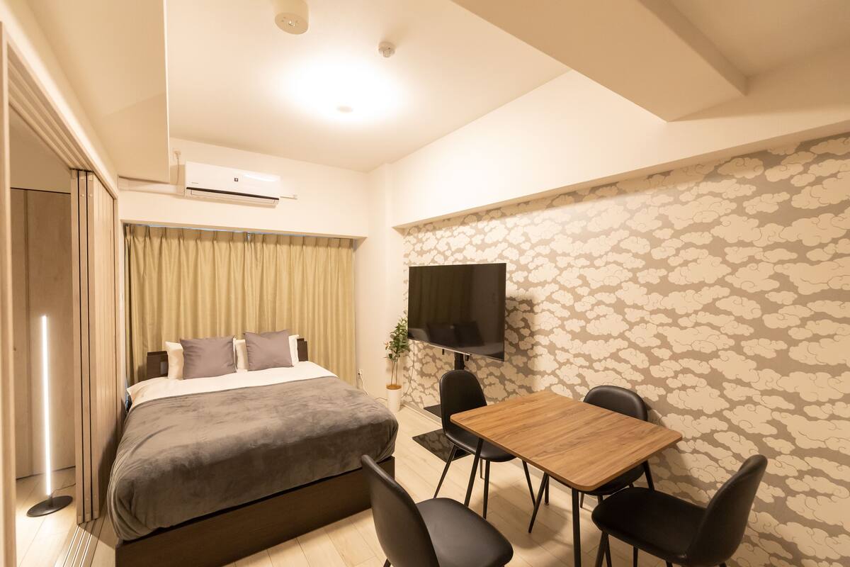 A hotel room with a bed, a TV, a dining table with chairs, and a patterned wall.