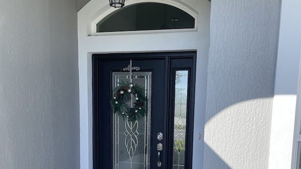 A black front door with a glass panel, a welcome wreath, and a small porch area with a doormat.