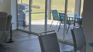 A modern living room with a glass table and chairs, a sofa, and a view of a patio with outdoor furniture and a barbecue grill.