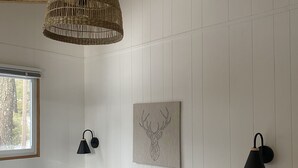 Ein Schlafzimmer mit Holzbalkendecke, einer gewebten Pendelleuchte, einer Wandleuchte, einer mit Rahmen versehenen Hirschdarstellung als Kunstwerk, einem Bett mit gestreifter Bettwäsche und einem kleinen hölzernen Hocker.