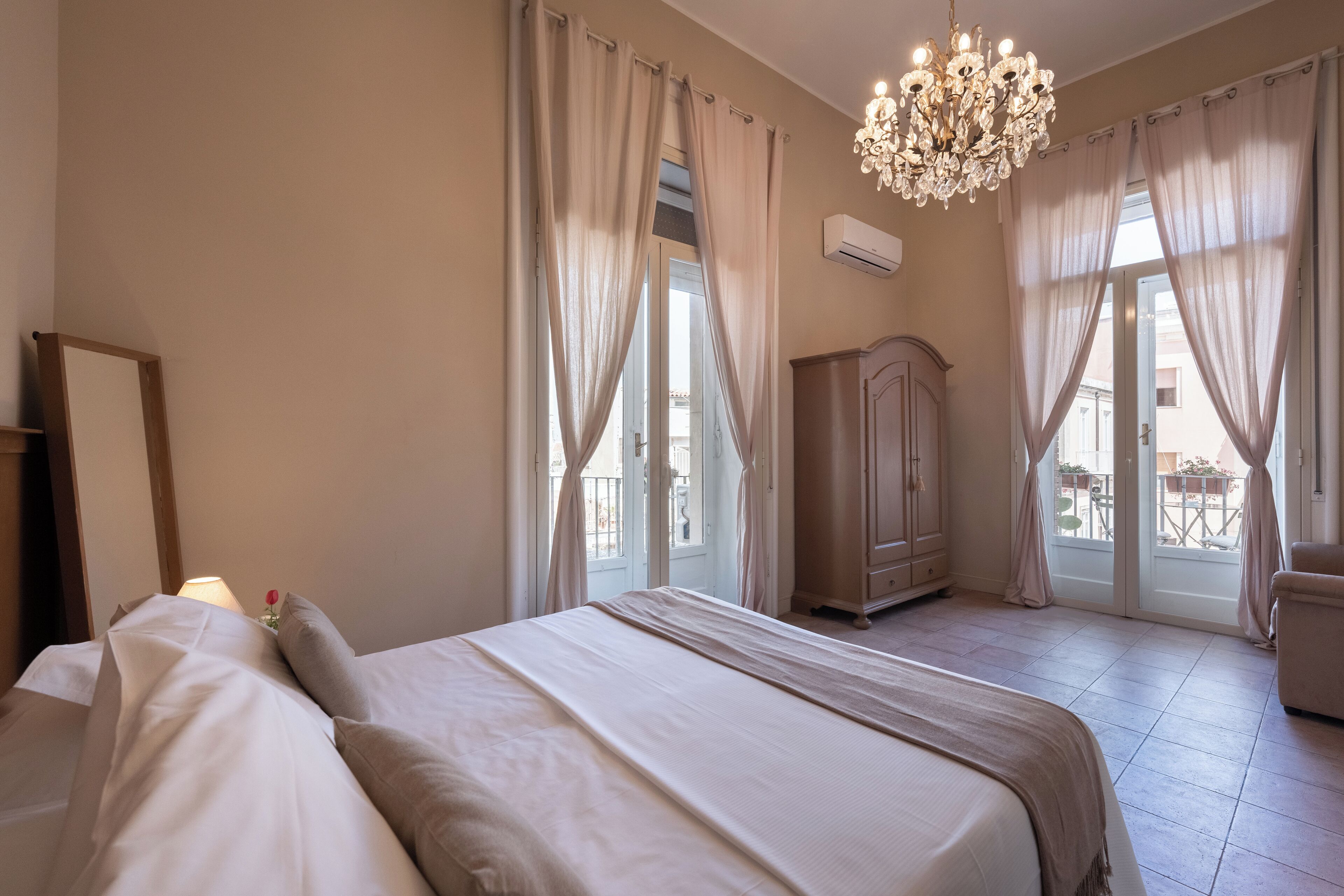 Un dormitorio con cama, cómoda, araña y ventanas grandes con cortinas.