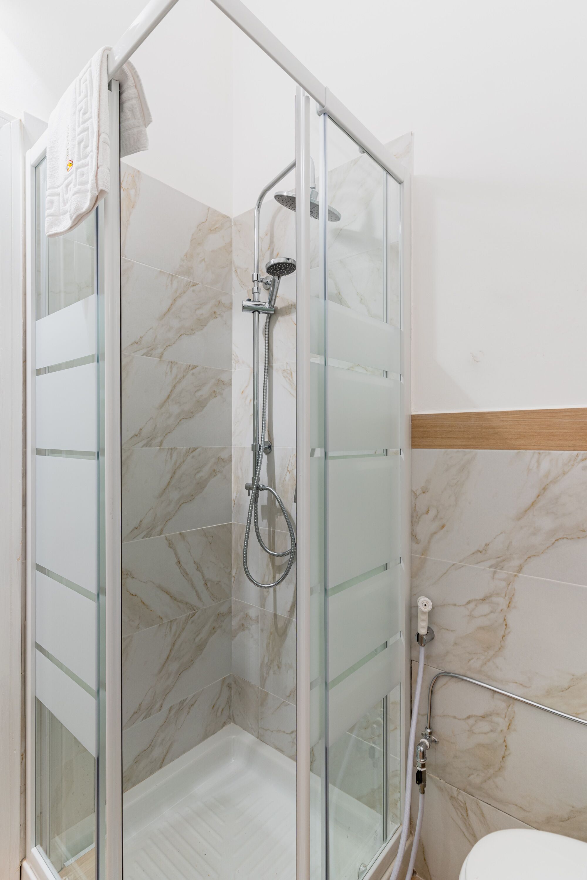 A glass-enclosed shower with a handheld showerhead, a glass door, and a marble-tiled wall.