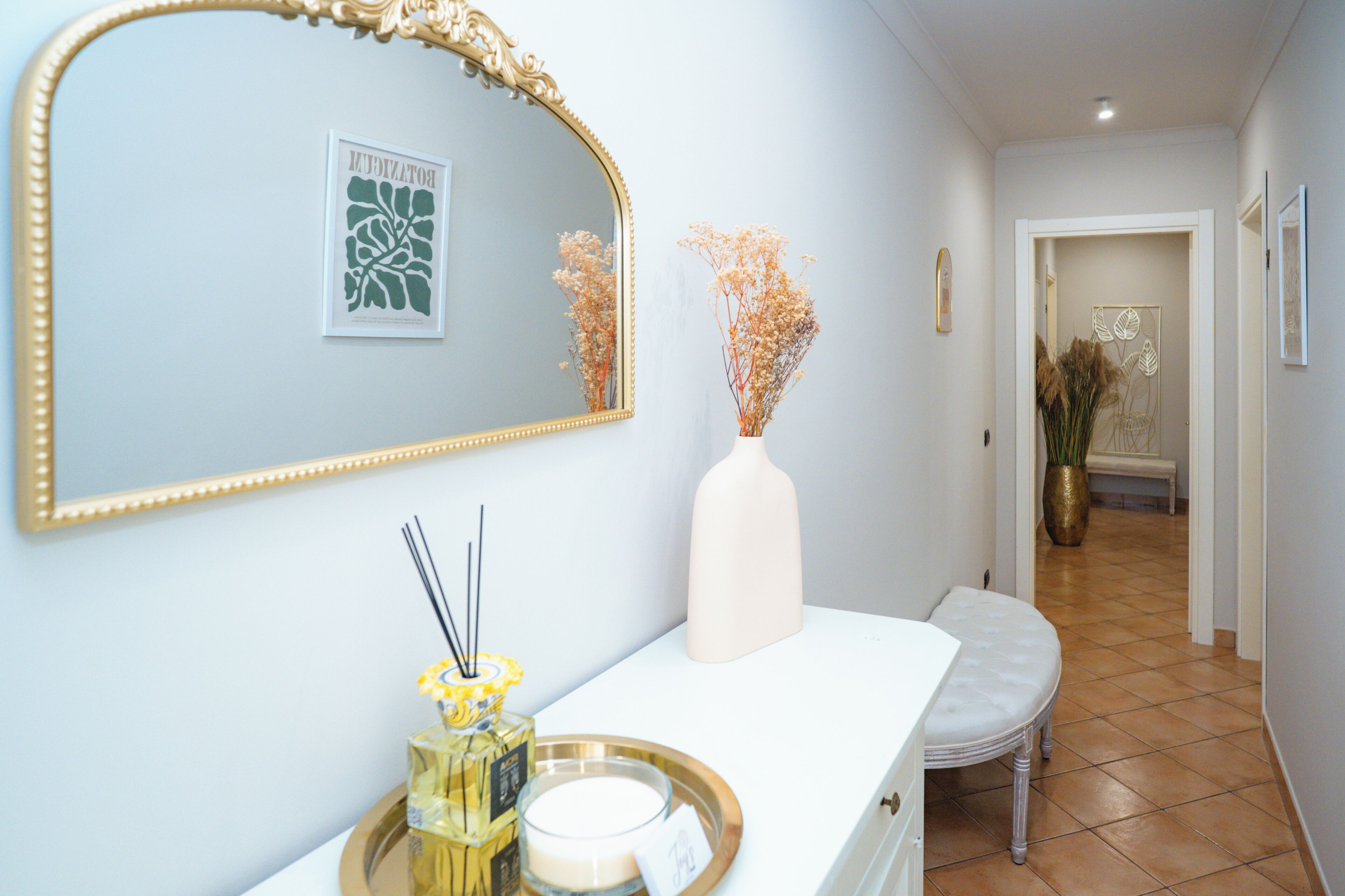 Un couloir avec un banc blanc, un miroir à cadre doré et un vase contenant des fleurs séchées.