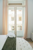 Una habitación de hotel con una cama, un tapete verde, un cabecero blanco, una ventana con cortinas y vistas a un balcón con sillas.
