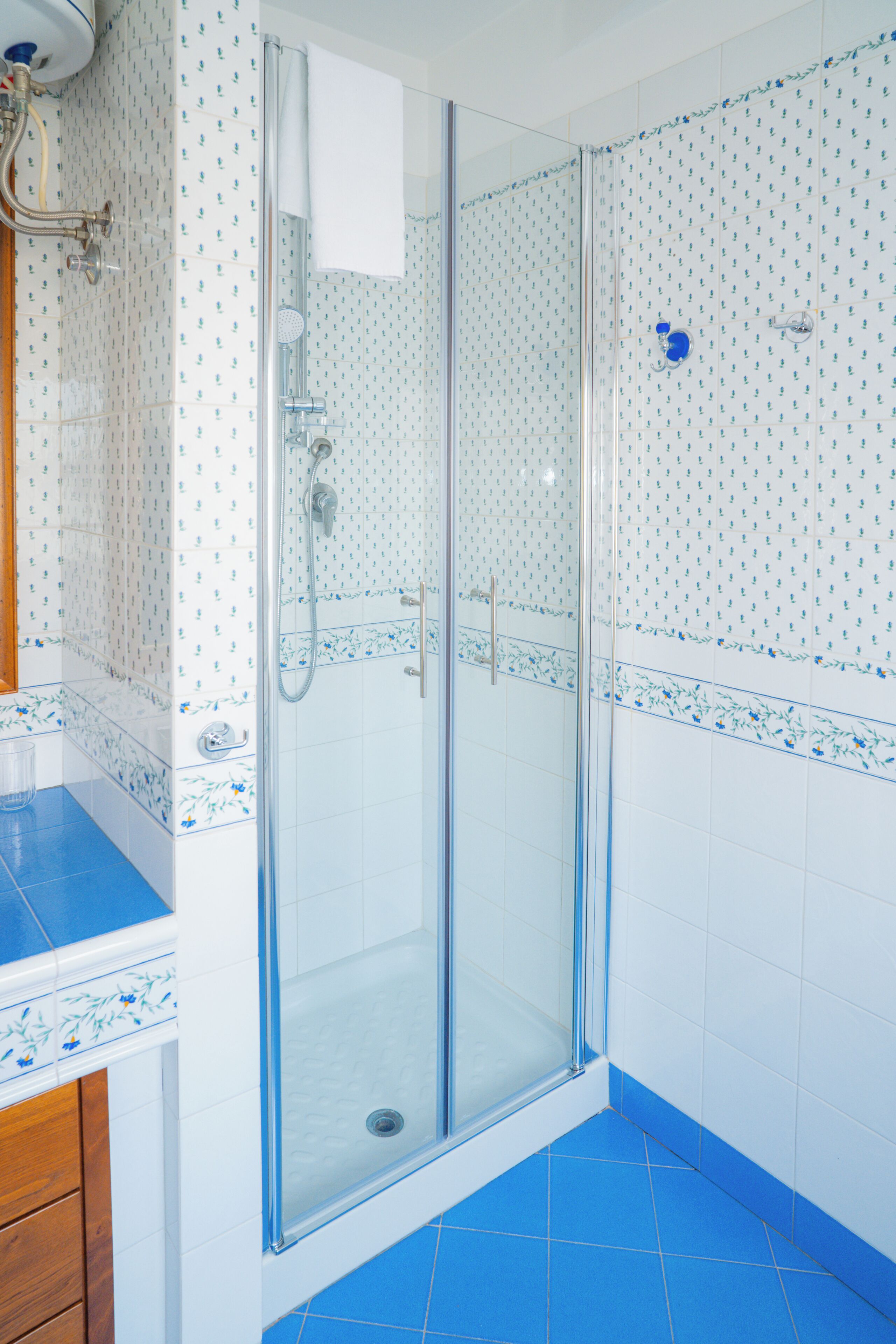 Eine Dusche mit Glaswand, ein weißes Handtuch am Türhänger, ein blauer Fliesenboden und wandbunte Fliesen.