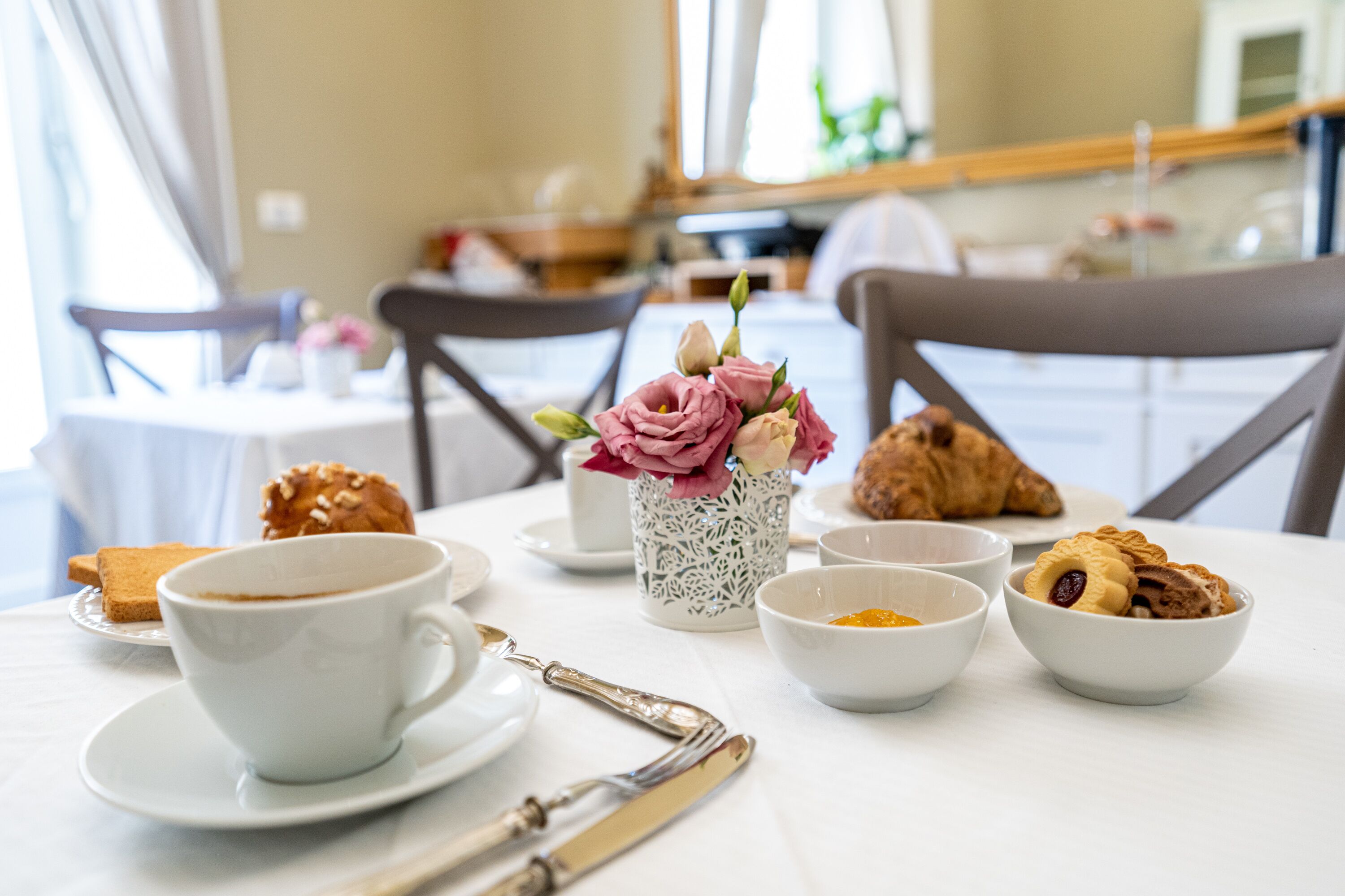 A breakfast setting with a cup, saucer, croissant, muffin, and a vase with flowers on a table.