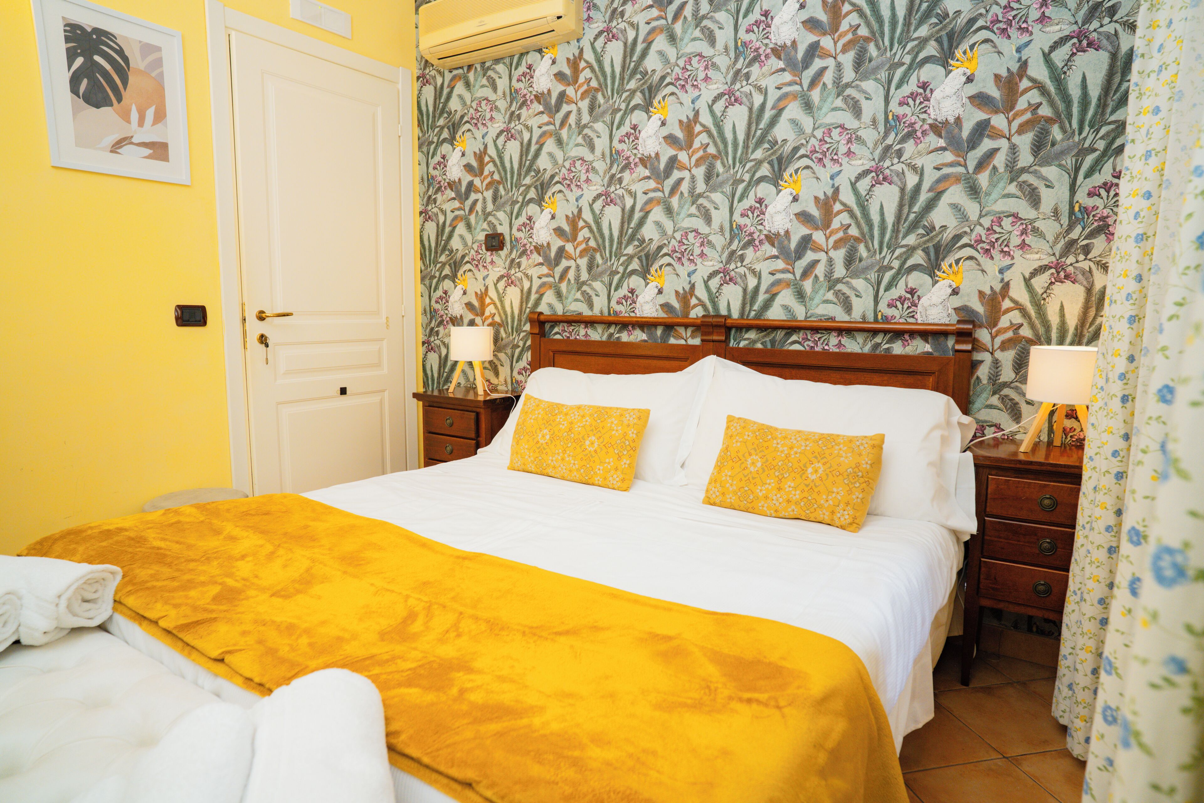 Una habitación de hotel con una cama, cabecera de madera, papel tapiz floral, colcha amarilla y puerta blanca.