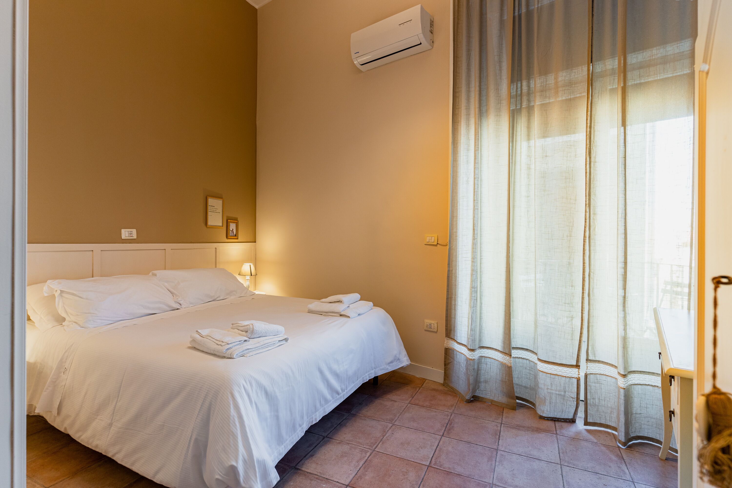 A hotel room with a bed, a window with sheer curtains, an air conditioning unit, and a chair.