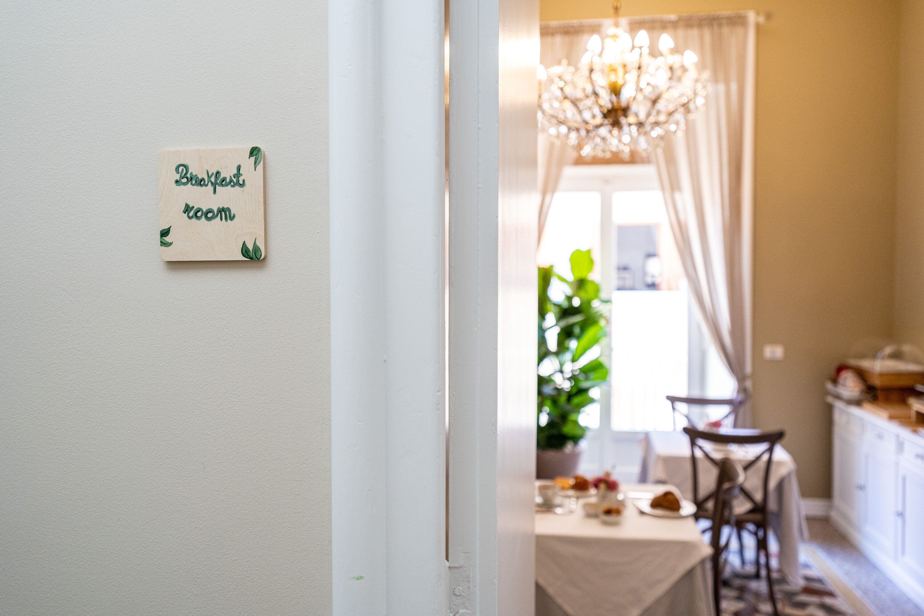 Un salón de desayunos con un cartel que dice 'Breakfast room', una araña, una mesa con sillas y una planta en maceta.