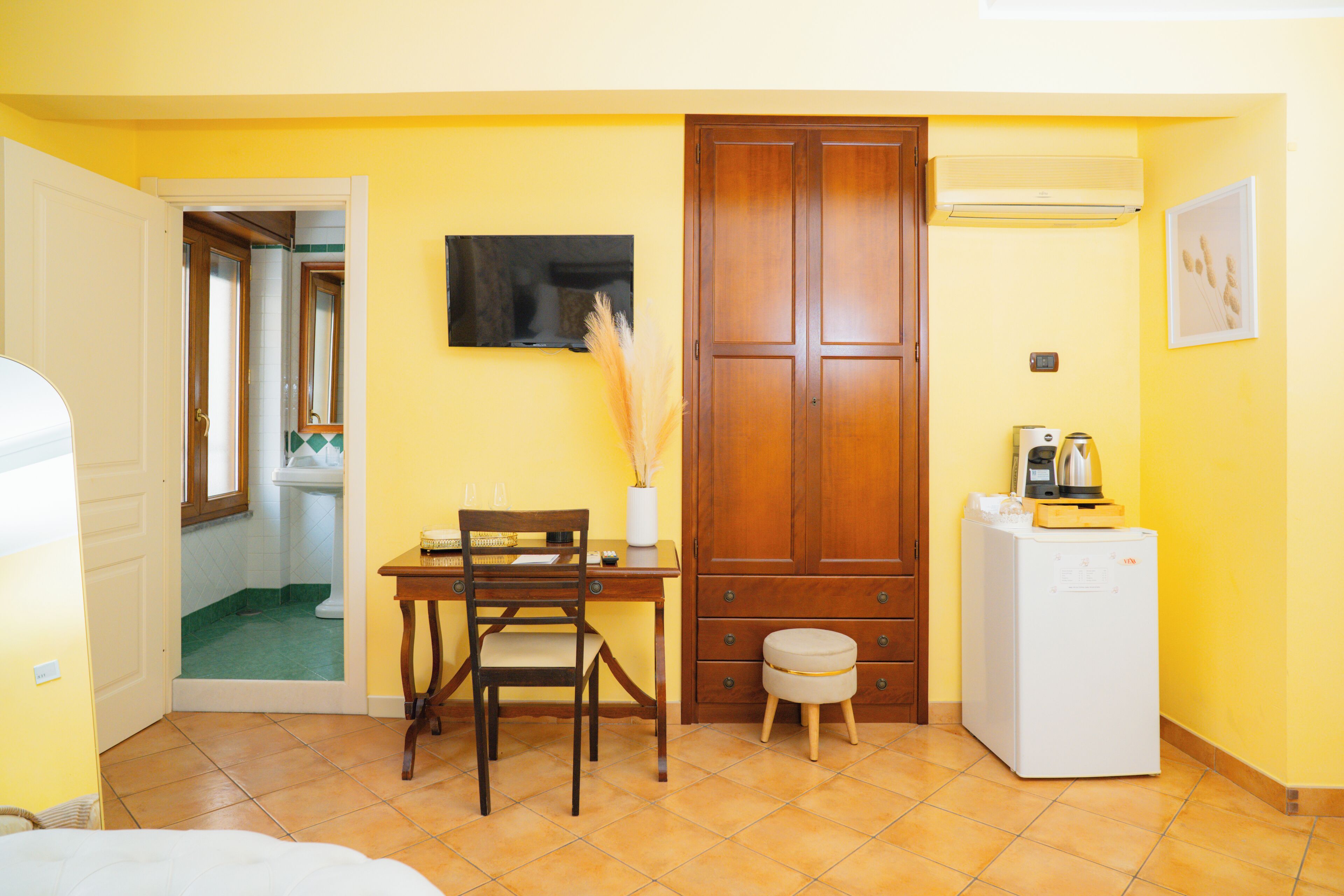 A compact room with a wooden door, a small table with chairs, a white refrigerator, and a television mounted on the wall.