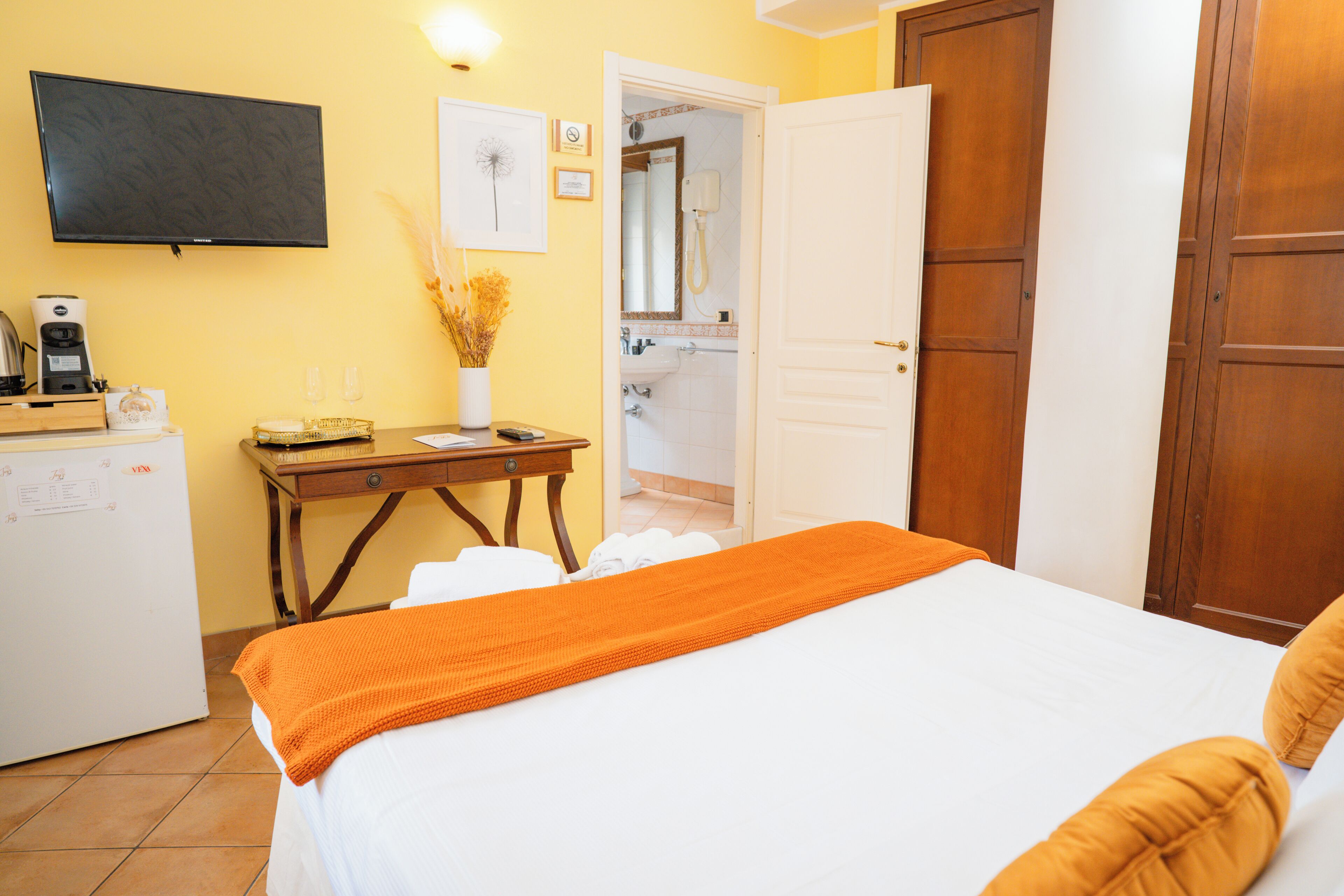 Habitación de hotel con cama, escritorio con cafetera, televisión y baño visible a través de una puerta abierta.